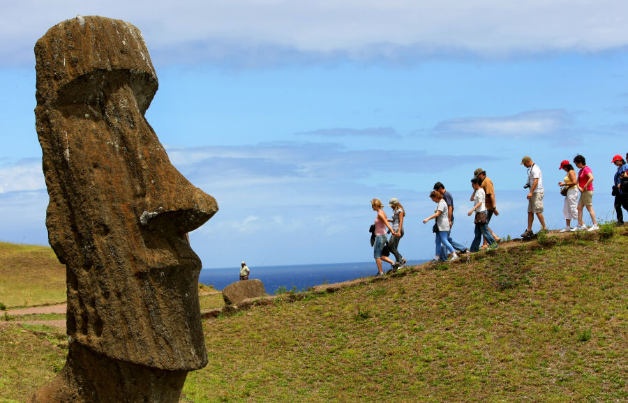 Należąca do Chile Wyspa Wielkanocna - zielone wzgórze, na pierwszym planie monolityczny posąg Moai, w tle grupa turystów zwiedzających wyspę