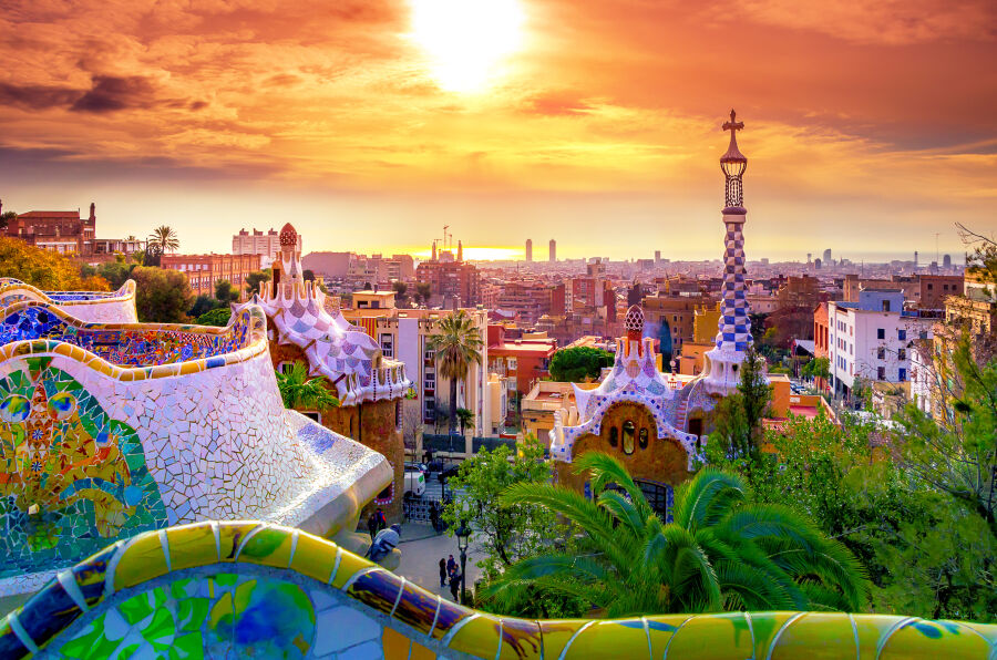 Widok ma miasto w promieniach zachodzącego słońca. Na pierwszym planie wieże i dachy budynków z charakterystyczną architekturą Gaudiego. 