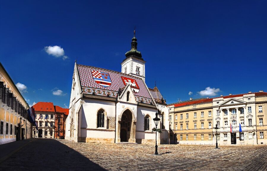 Plac św. Marka w Zagrzebiu. Centralnie posadowiony zabytkowy kościół z barwnym dachem.  