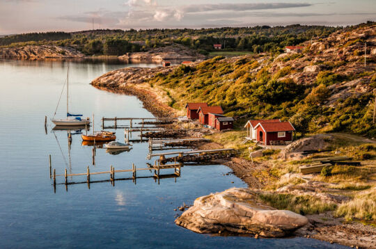 Zachodnie wybrzeże Szwecji. Skaliste wybrzeże i zatoka po lewej kilka małych kolorowych domków oraz drewniane przystanie z kilkoma niewielkimi łodziami 